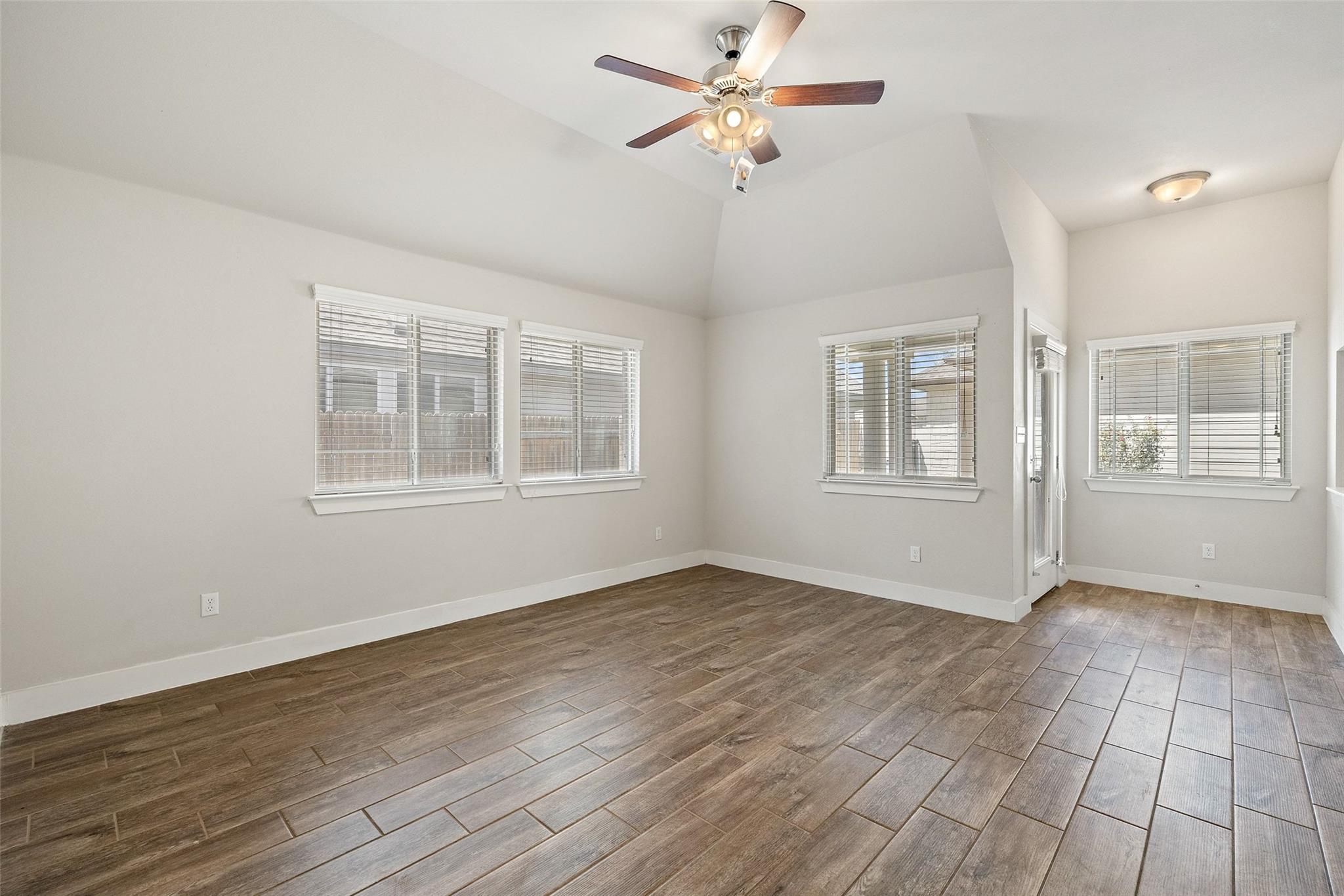 Empty vaulted room with a ceiling fan, large windows, and brown wood-look tile flooring.
