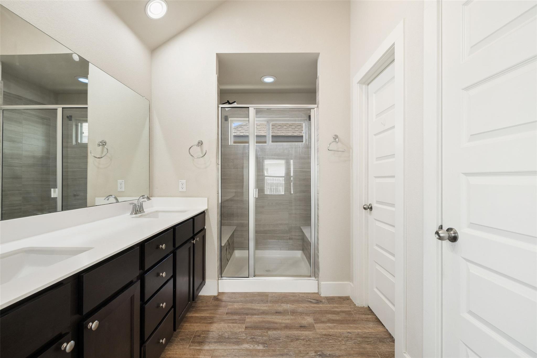 Bathroom with a dark vanity, large mirror, glass-enclosed tiled shower, and wood-look tile floor.
