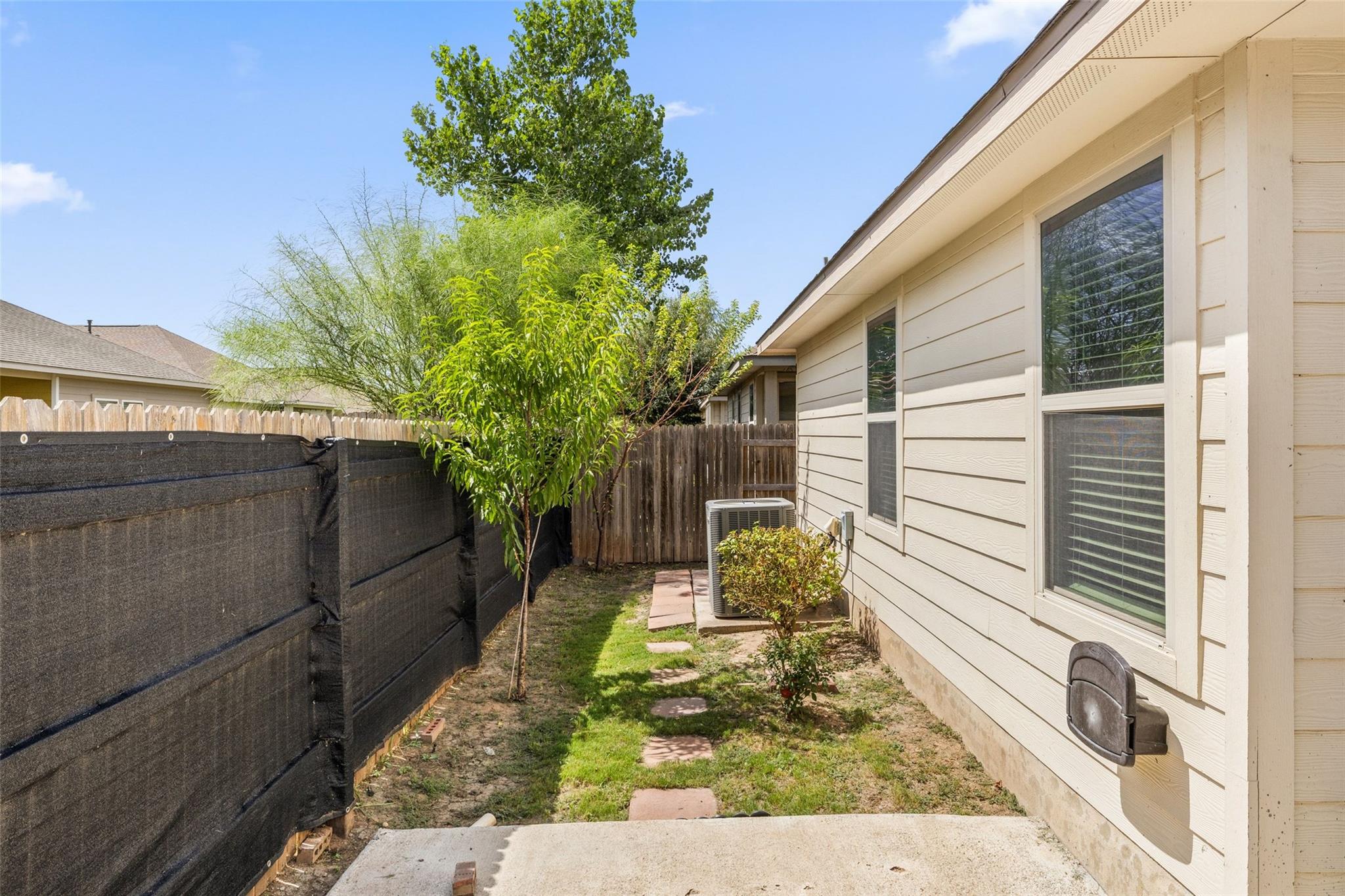 Side yard with a stepping-stone path, trees, and a fenced boundary beside a beige house.