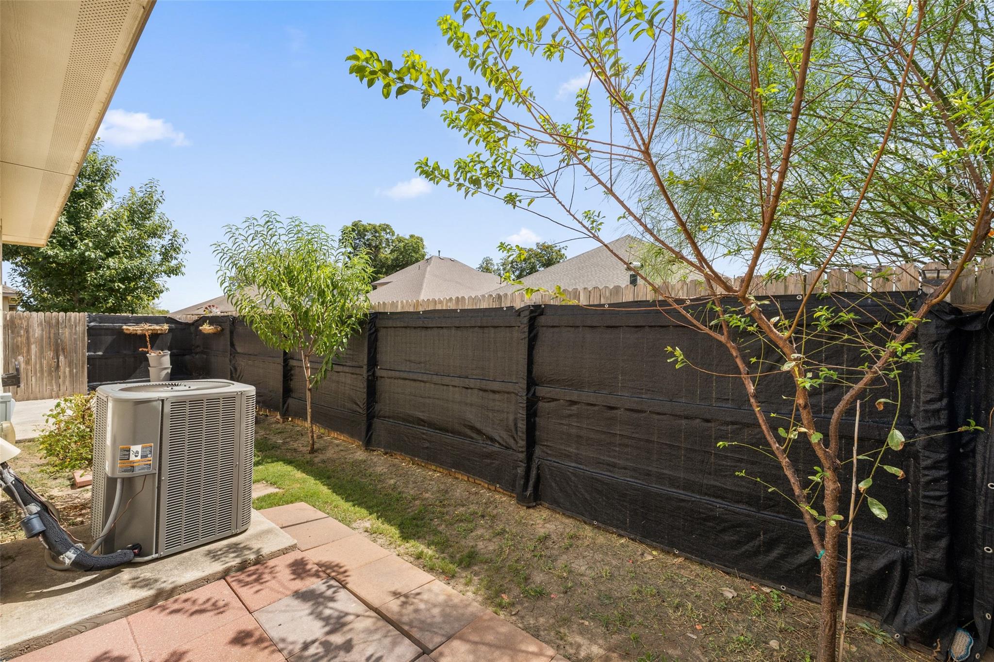 Backyard with a paved patio, central air-conditioning unit, young trees, and black privacy screening on the fence.
