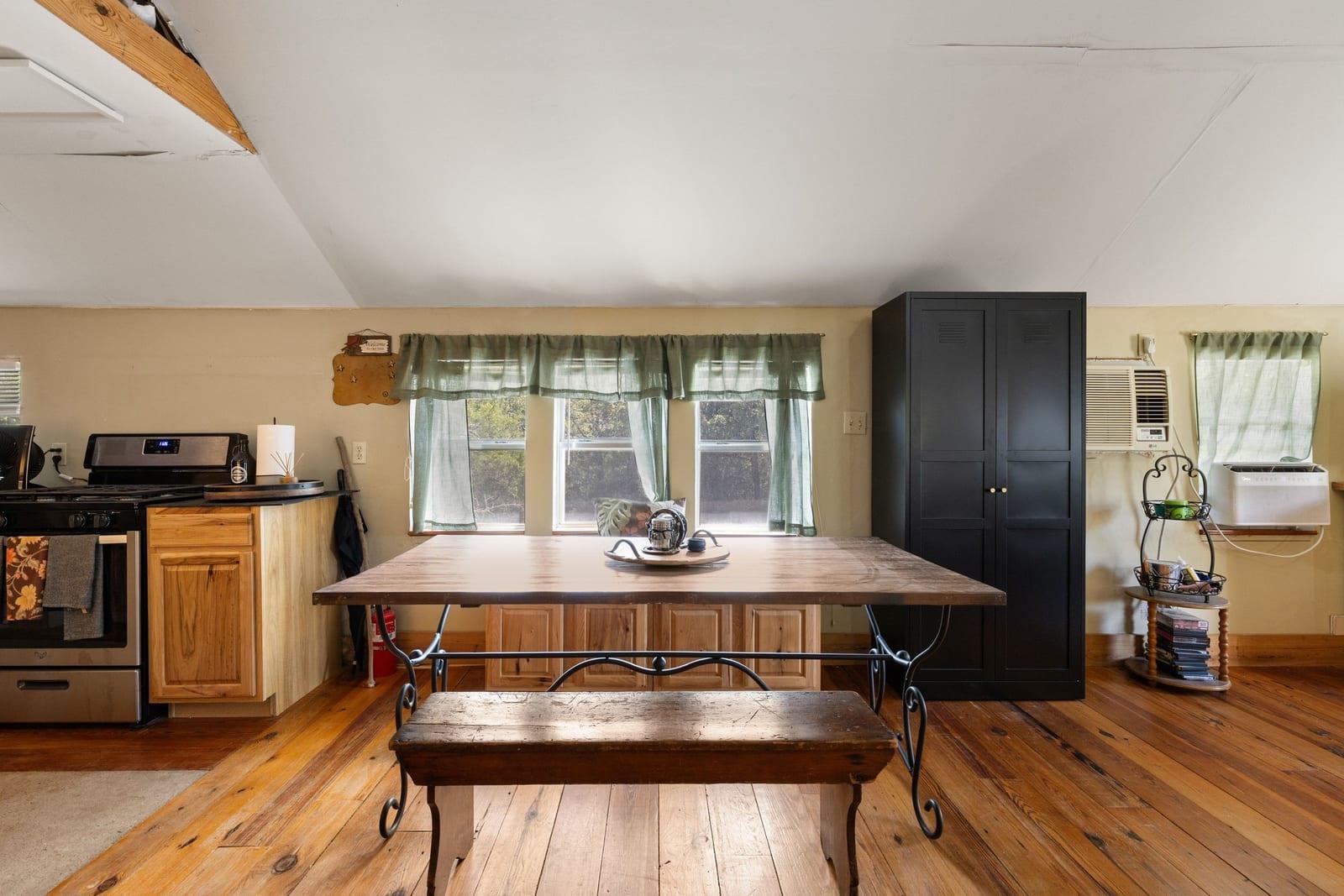 Kitchen dining area with a long wooden table, green-curtained windows, and hardwood floors.