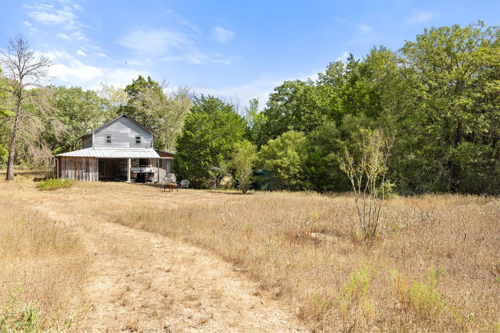 Two-story house with a covered porch in a wooded clearing, reached by a dirt path.