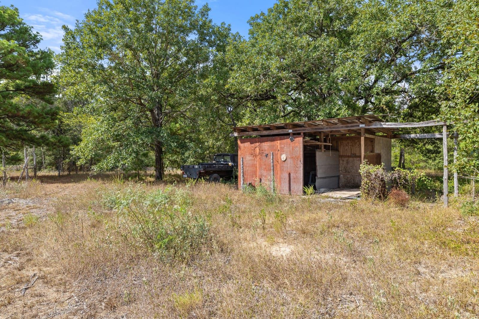 Wooden shed and open carport in a dry, tree-shaded clearing, with a black pickup truck nearby.