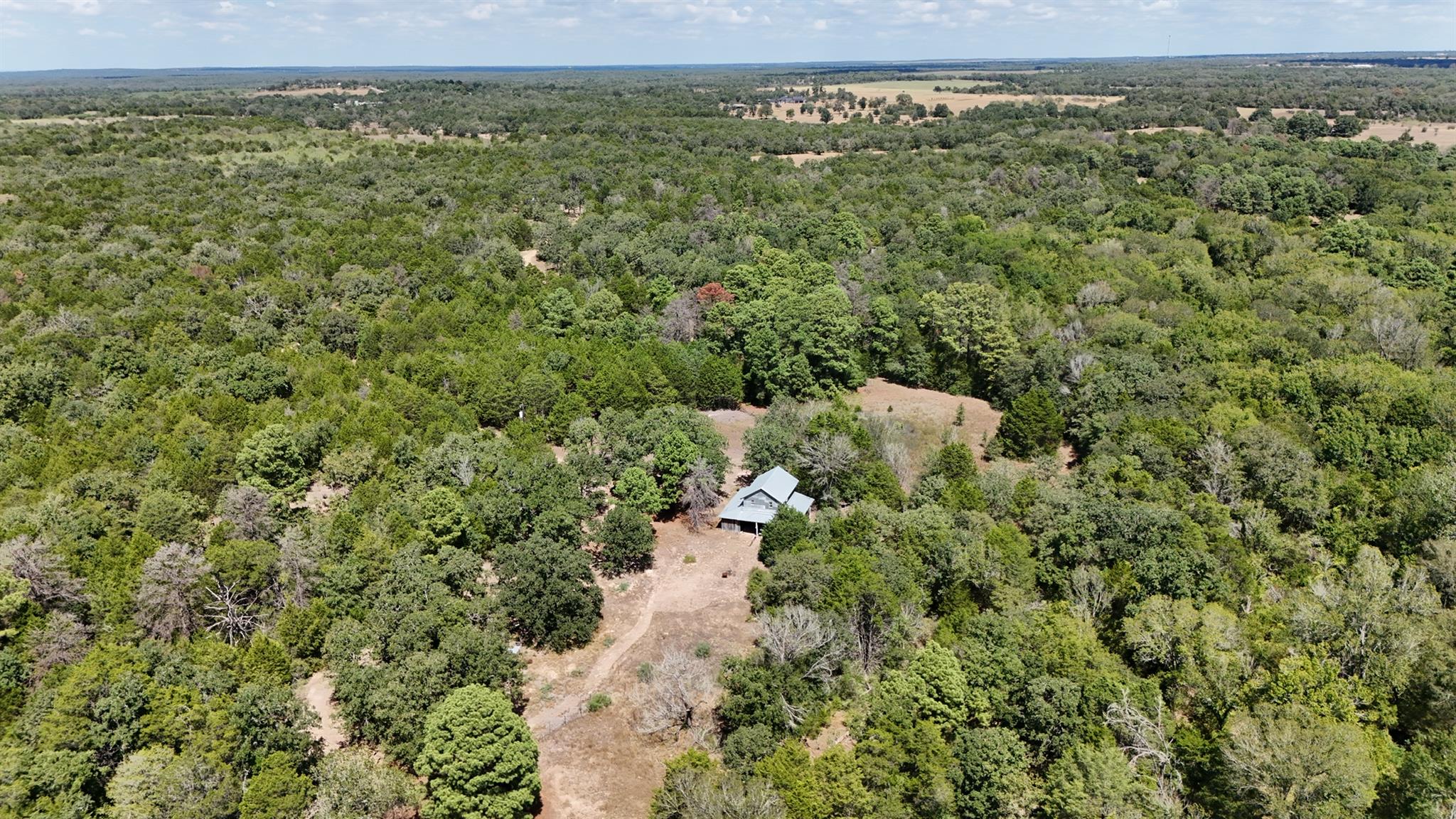 Aerial view of a house surrounded by dense woods, with a cleared yard and access track.