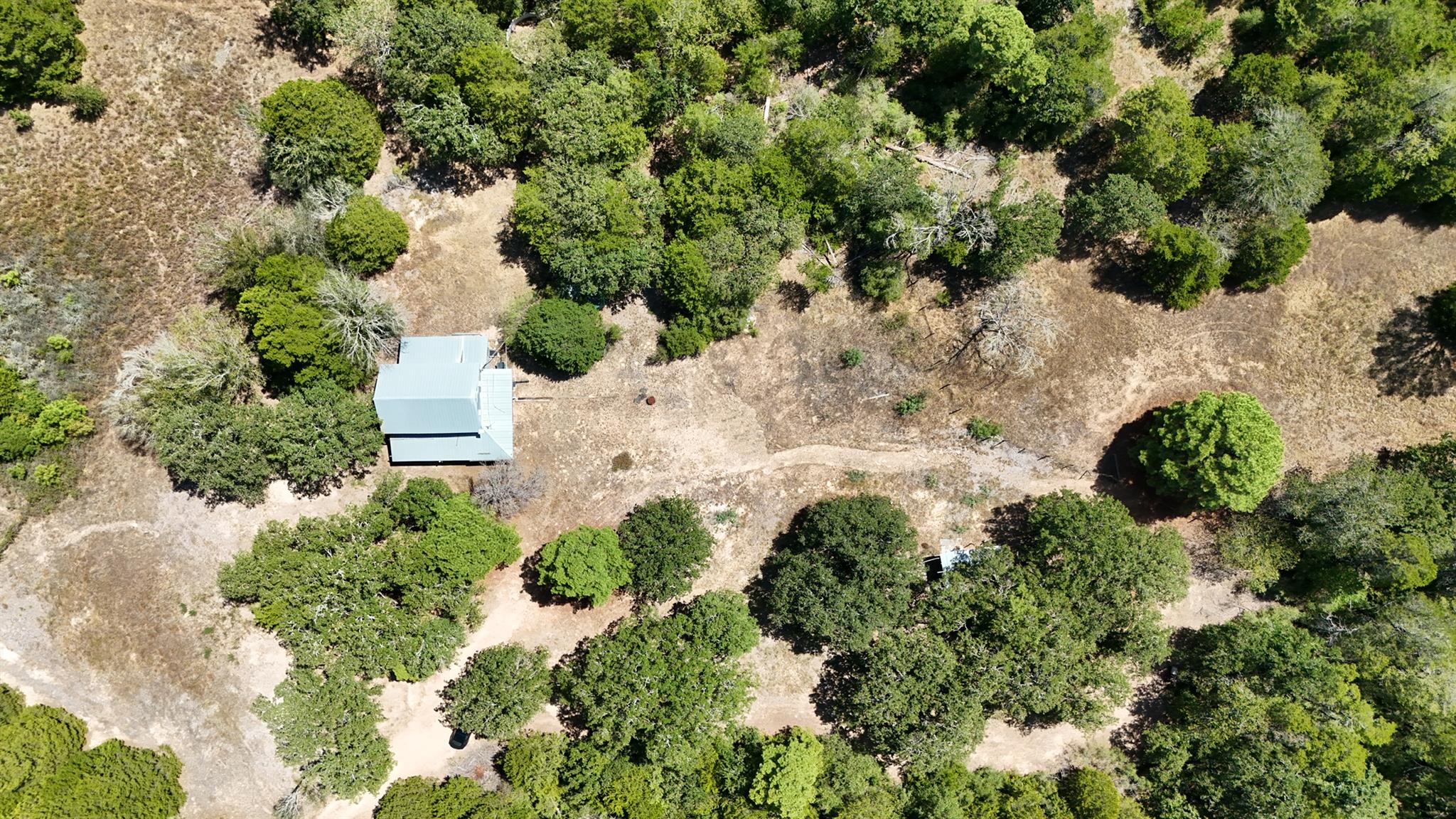 Aerial view of a wooded property with a metal-roof outbuilding and open dry clearing.