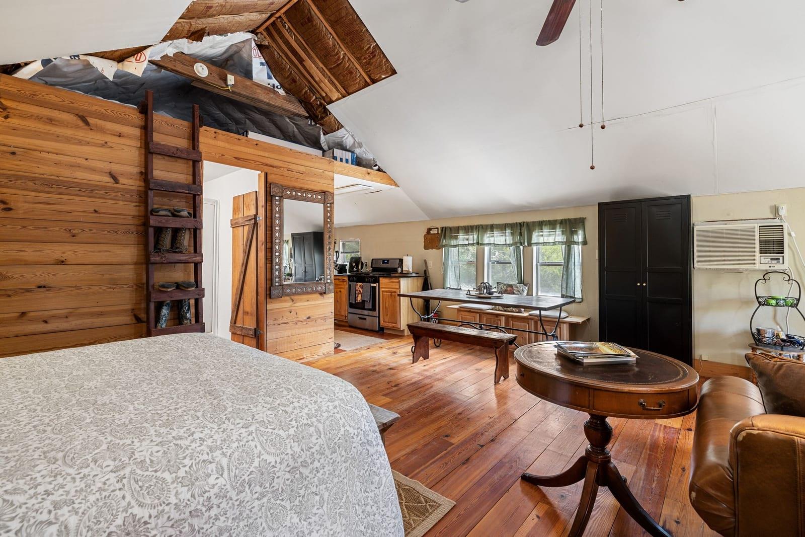 Open-plan bedroom with a wood-paneled loft wall, exposed rafters, compact kitchen, and hardwood floors.