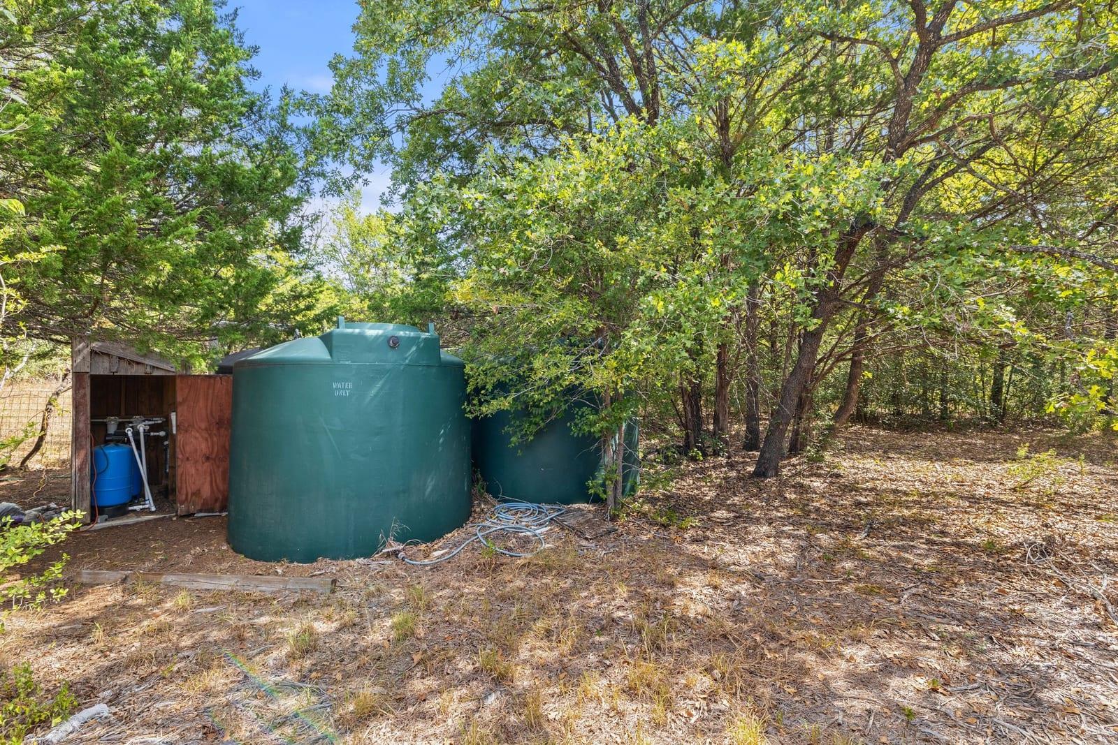 Green water storage tanks and a pump shed beneath trees in a wooded yard.