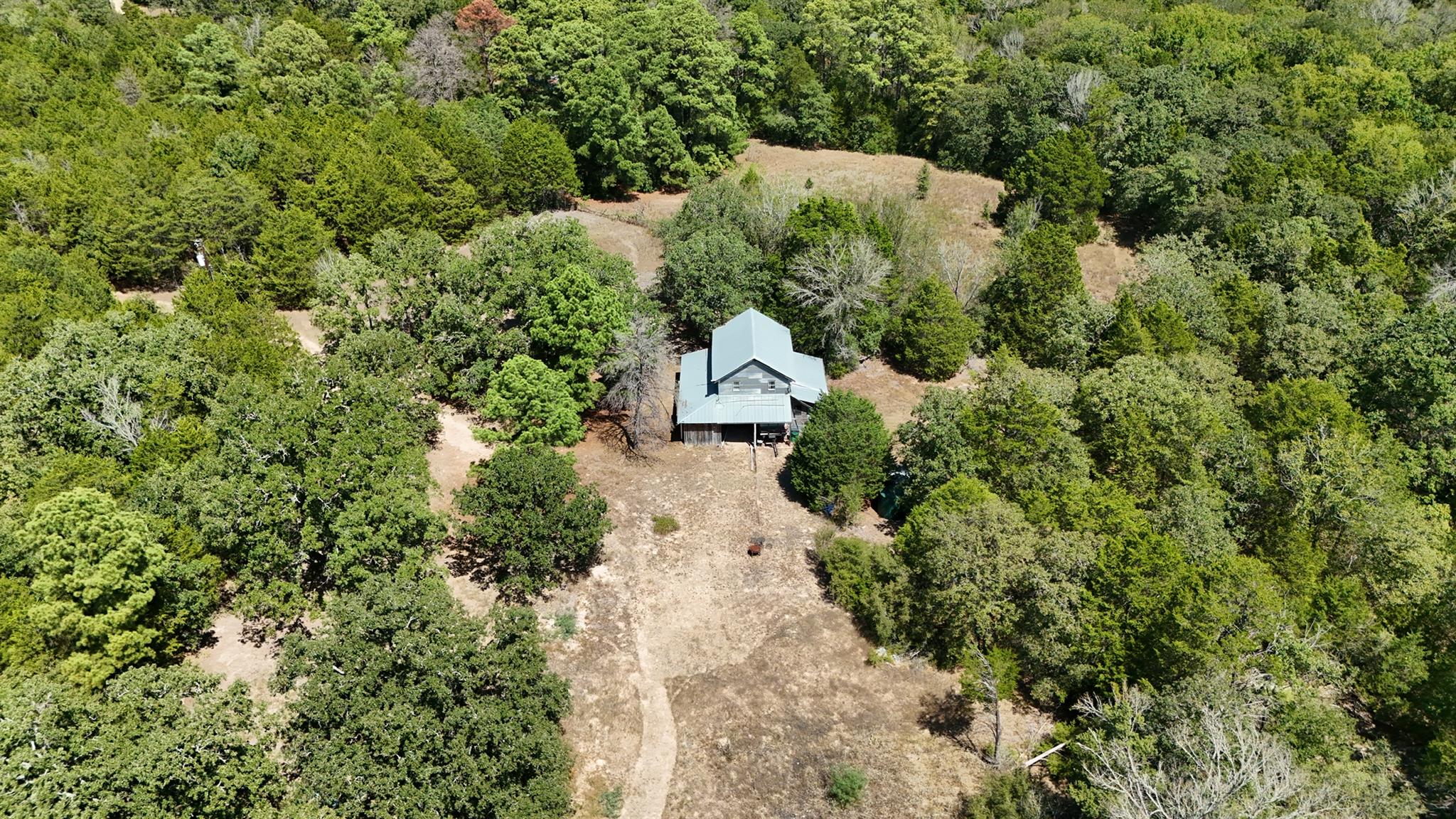 Aerial view of a metal-roofed house in a wooded clearing, with a dirt driveway.