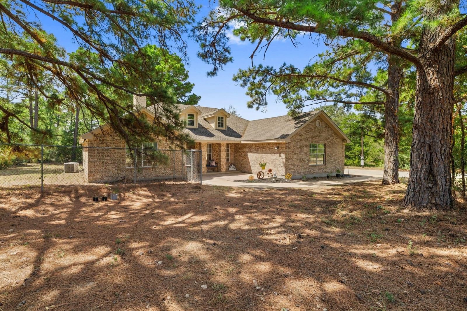 Brick house with dormer windows and a paved patio, framed by tall pine trees.