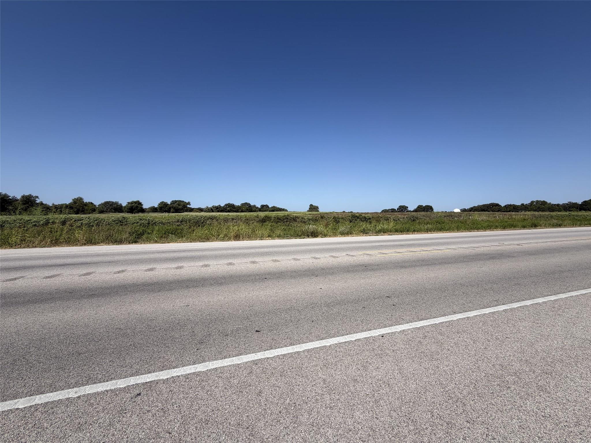 Vacant grassy land across a highway, with a distant tree line and clear blue sky.