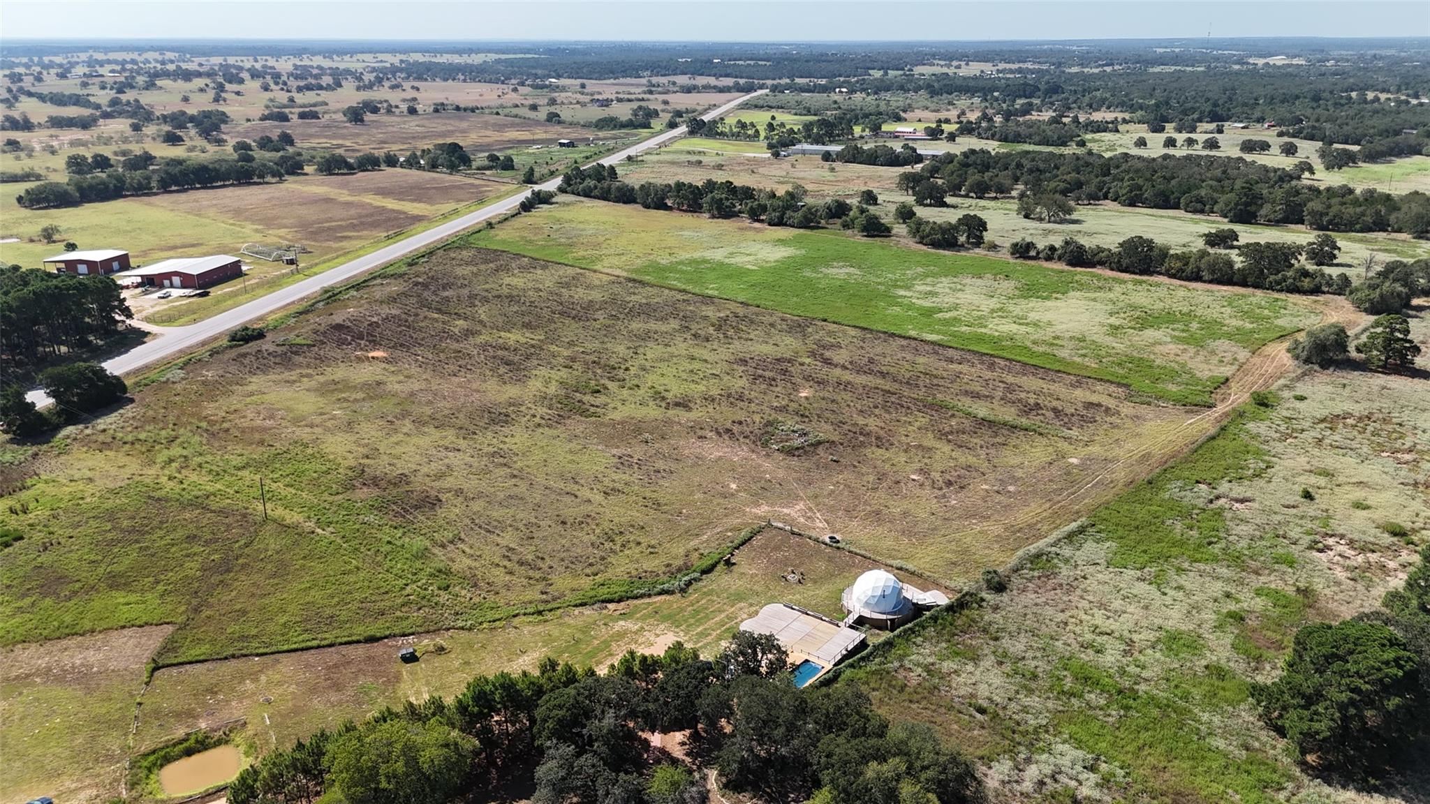 Aerial view of open rural land beside a highway, with a small pond and fields in the distance.