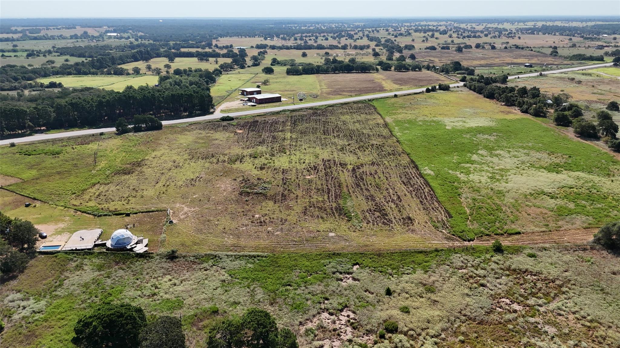Aerial view of rural acreage with fields, highway, outbuildings, and a geodesic dome beside a small pool.