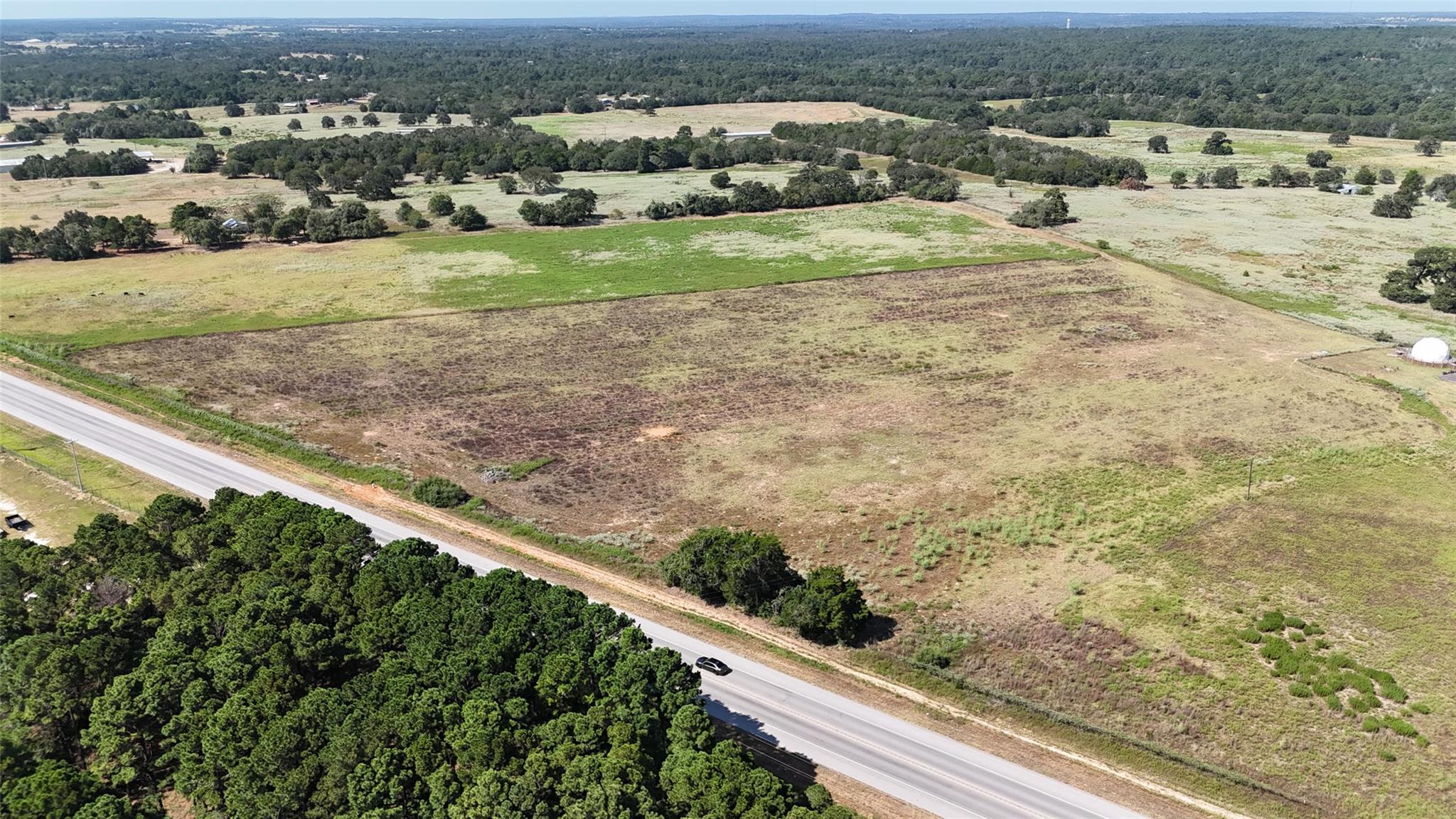 Aerial view of open pastureland bordering a highway, with scattered trees and woodland beyond.