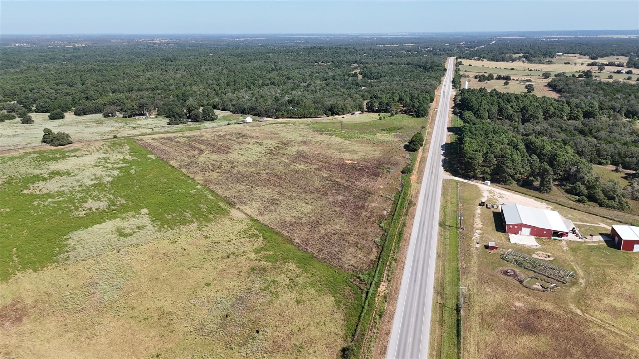 Aerial view of open rural land beside a highway, with woods and farm buildings nearby.