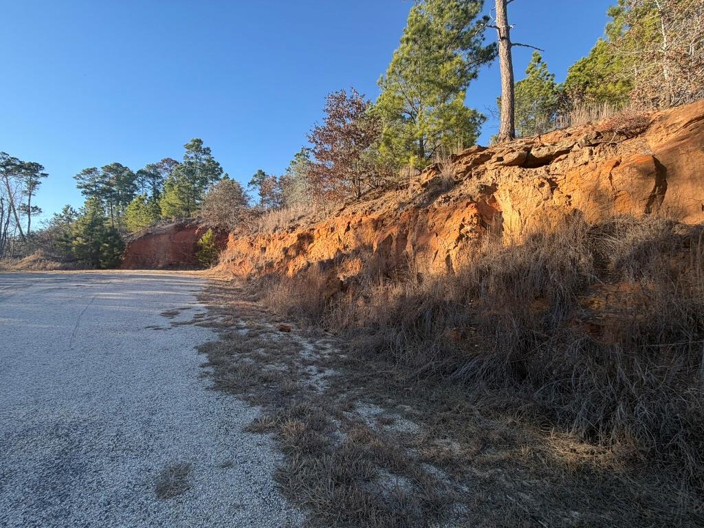 Gravel road beside a lot with a tall red rock embankment and trees.