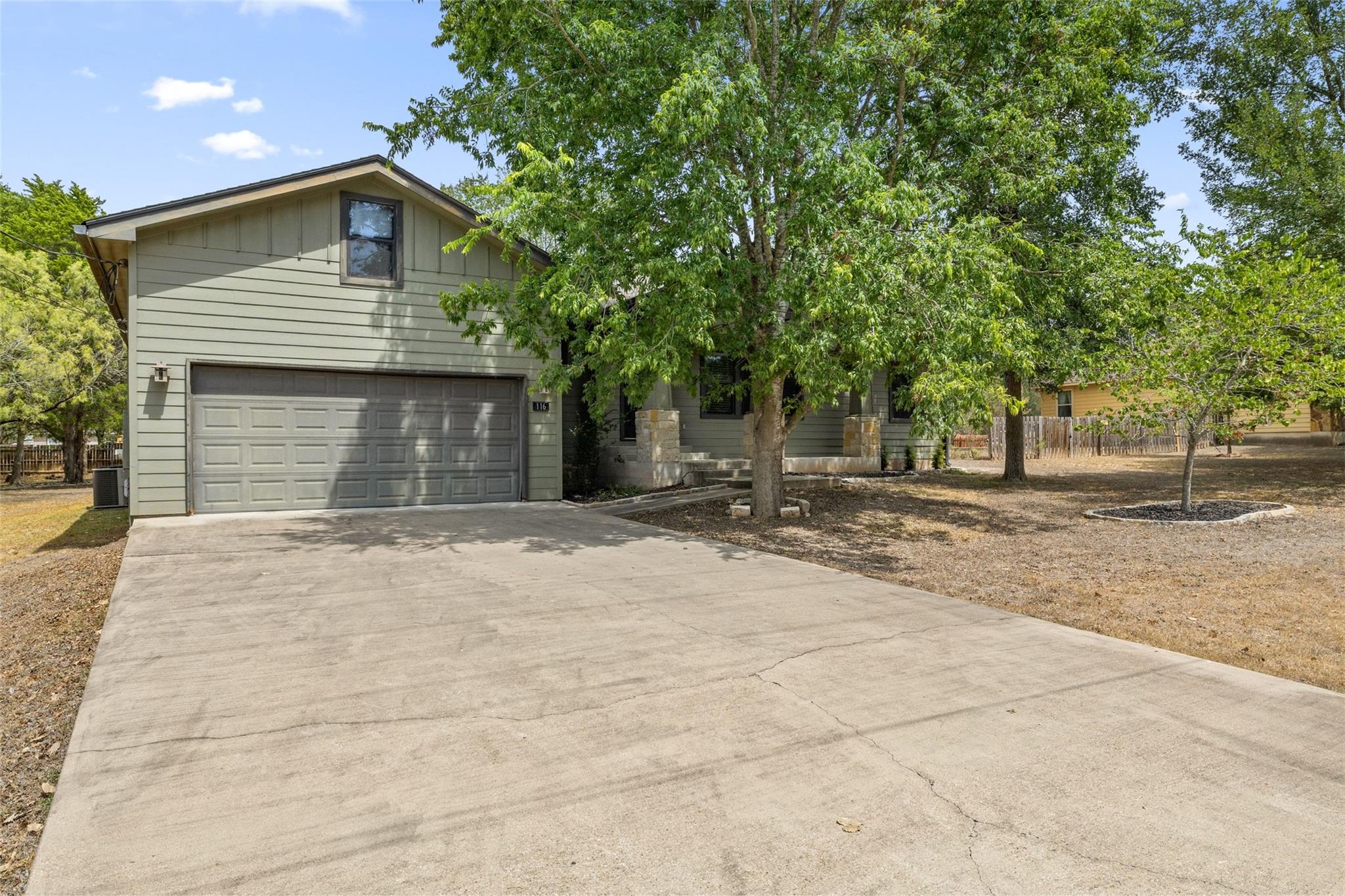Two-story gray-green house with a wide garage, broad driveway, and leafy trees shading the front porch.