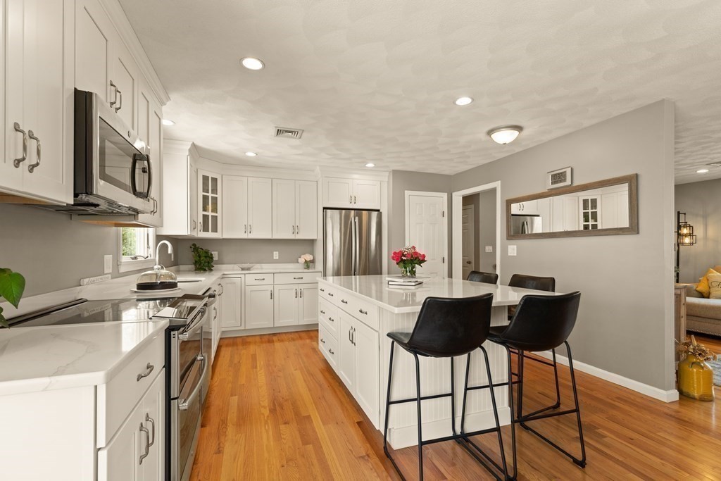 Kitchen with a white island, black bar stools, stainless steel appliances, and warm hardwood floors.