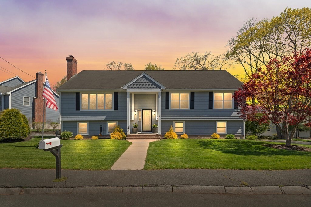 Blue-gray split-level house at dusk with a front walkway, lit windows, and autumn trees.