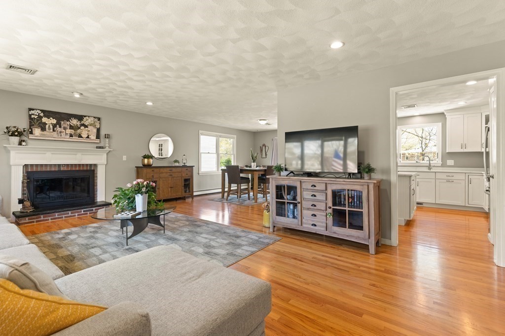 Furnished living room with a white fireplace, hardwood floors, and an adjoining kitchen.