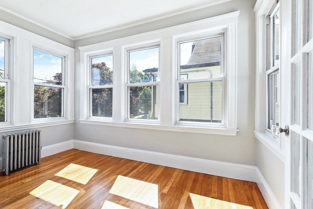 Empty room with polished hardwood floors, a cast-iron radiator, and large sunlit windows.