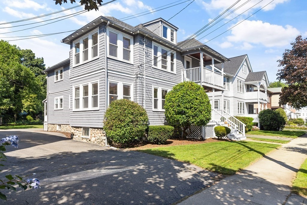 Gray shingle-sided home with white trim, a raised porch, front shrubs, and a driveway.