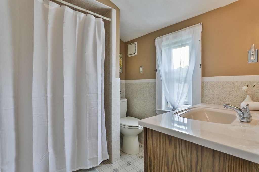 Bathroom with a white shower curtain, toilet, and wood-grain vanity beneath a bright window.