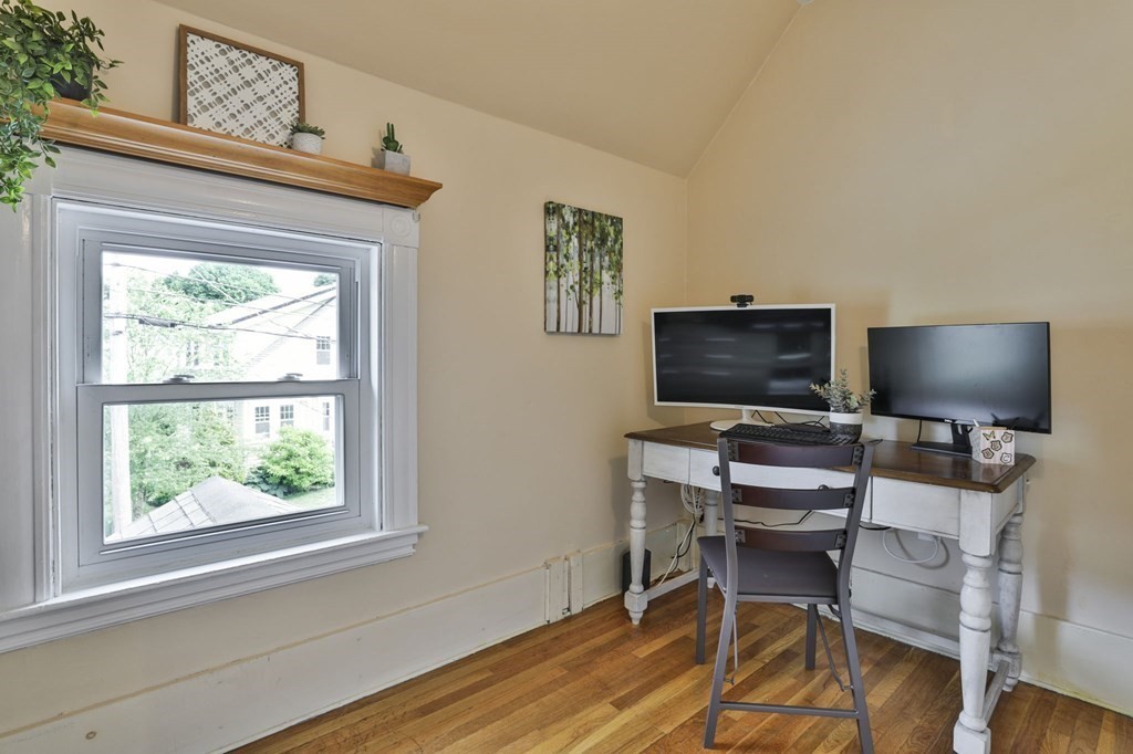 Home office with two monitors on a white desk beside a large window and hardwood floors.