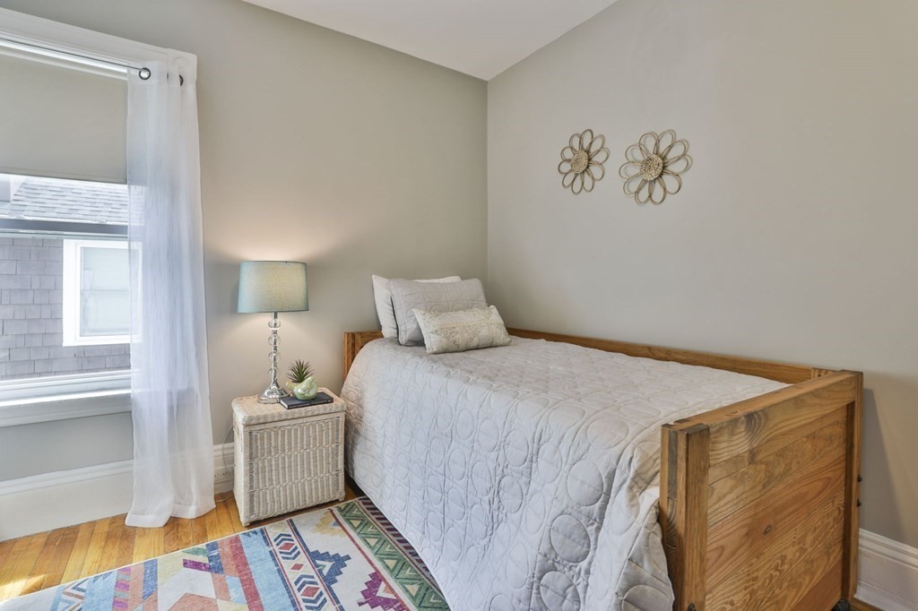 Bedroom with a wooden bed, white quilt, colorful geometric rug, and sheer curtains.