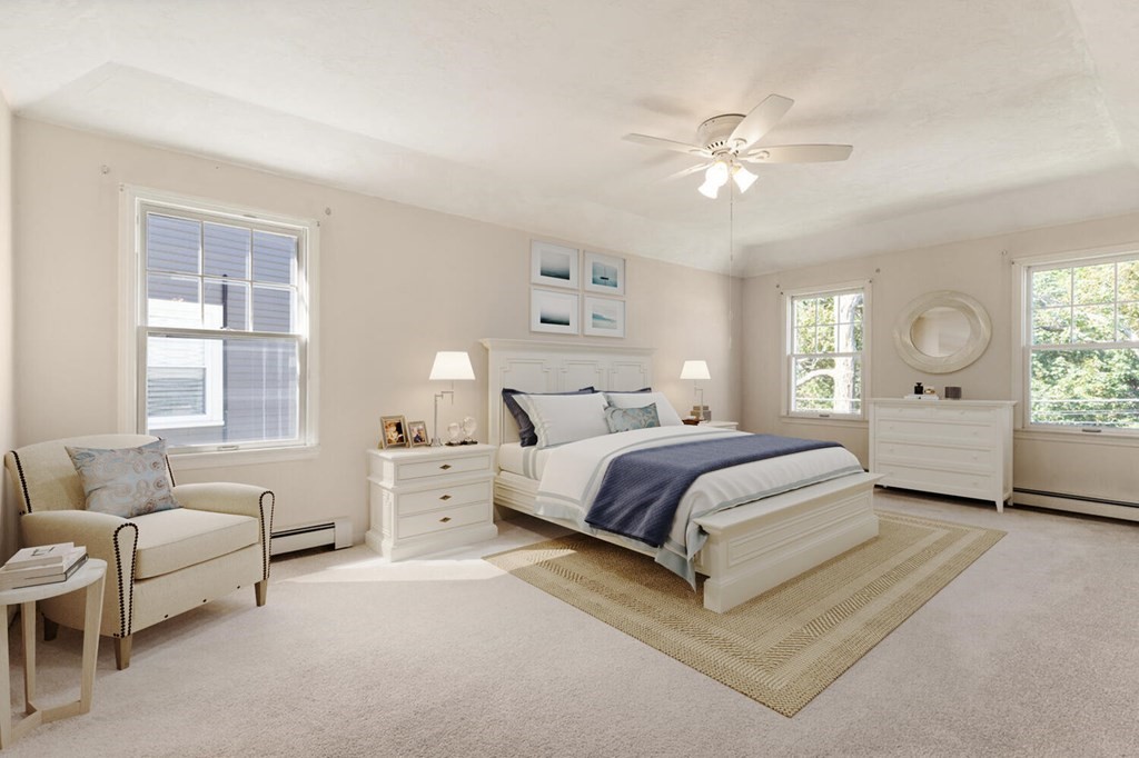 Furnished bedroom with a white bed, navy bedding, ceiling fan, and large windows.