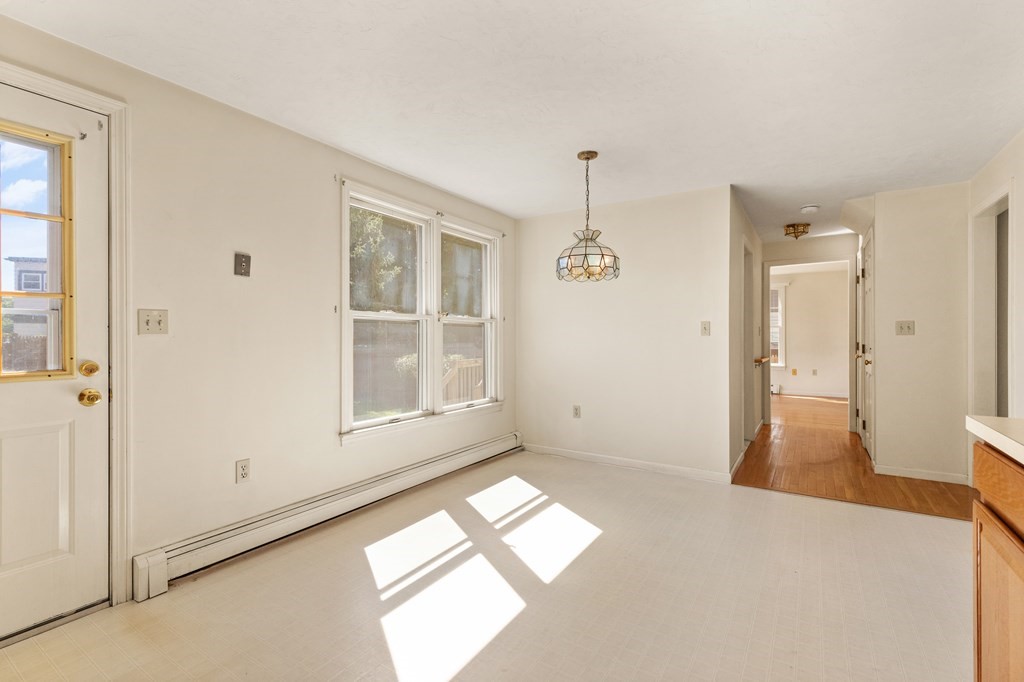 Empty dining area with pale tile floors, a stained-glass pendant, windows, and a hallway with hardwood floors.