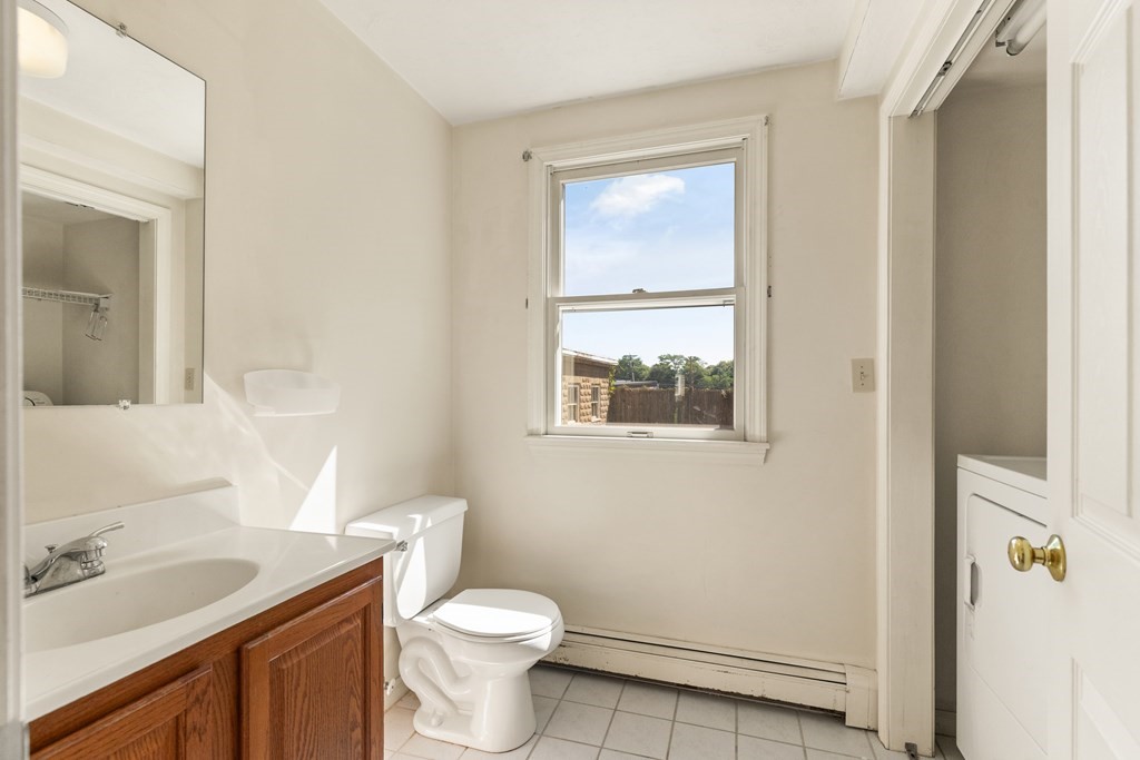 Vacant bathroom with a wood vanity, white toilet, tiled floor, and window overlooking a fenced yard.