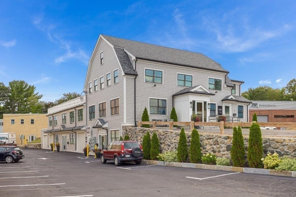 Gray multi-unit building with a covered entrance, stone retaining wall, landscaping, and parking lot.