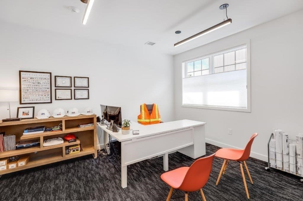 Furnished office with a white desk, coral guest chairs, wood shelving, and high windows.
