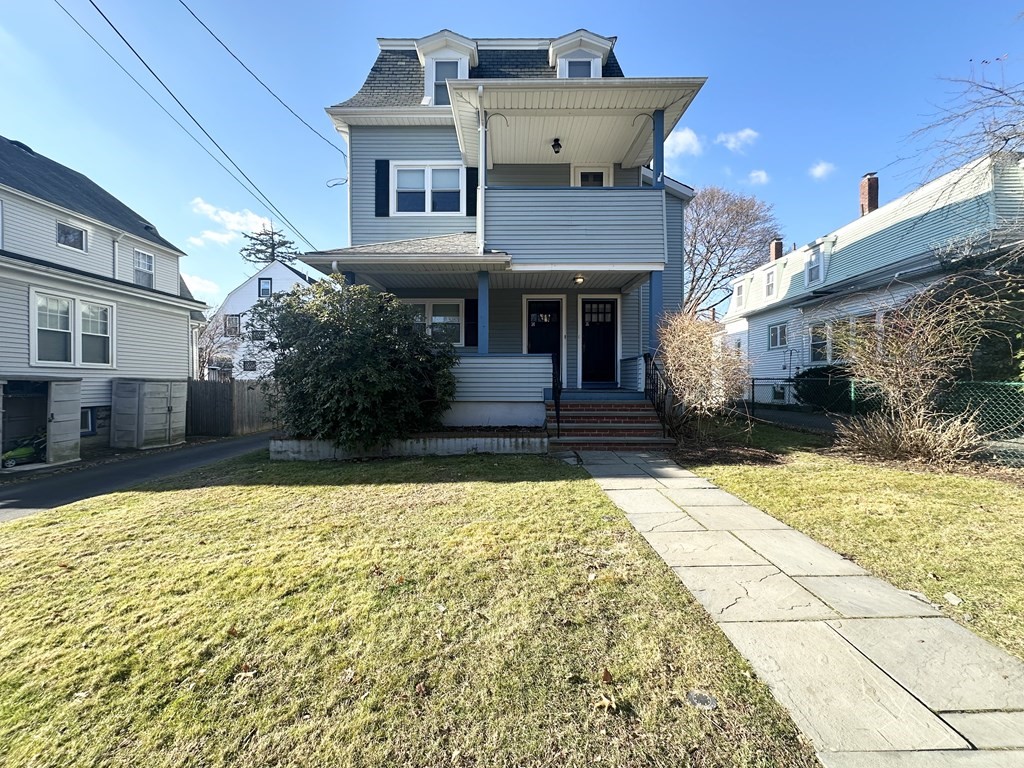 Blue-gray multi-story house with a covered porch, upper balcony, and concrete walkway across the lawn.
