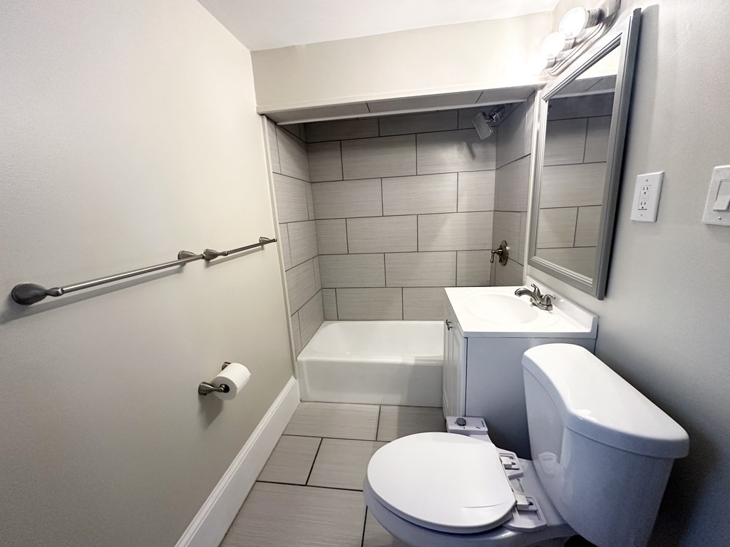 Bathroom with a gray tiled tub surround, white vanity, toilet, and framed mirror.