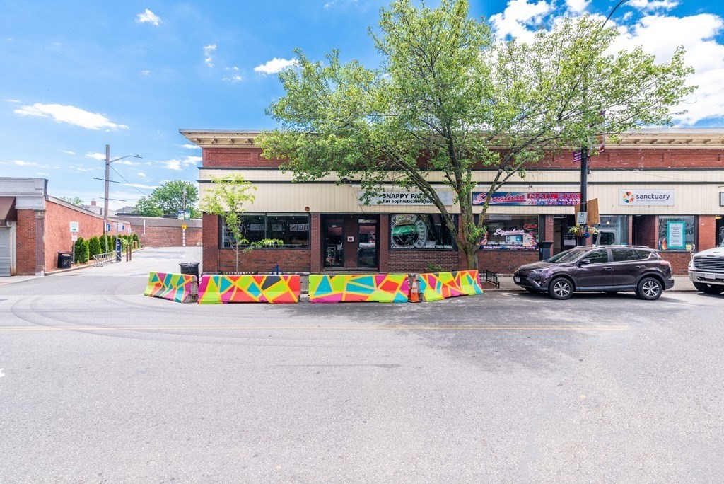 Commercial storefronts with a leafy tree and colorful barriers along the curb.
