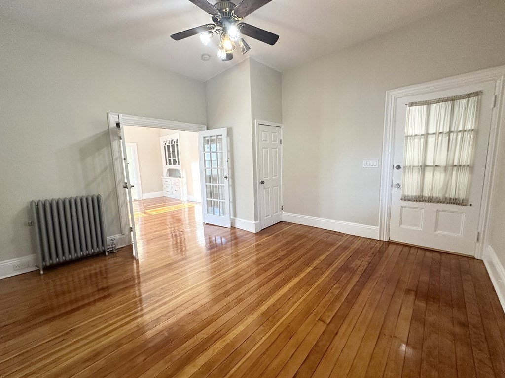 Empty room with polished hardwood floors, a ceiling fan, radiator, and French doors to an adjoining room.