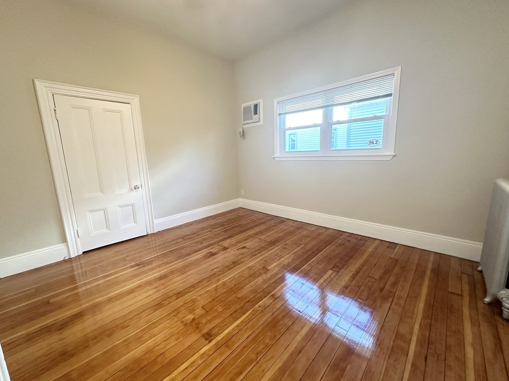 Empty room with polished hardwood floors, a window with blinds, and a wall-mounted air conditioner.