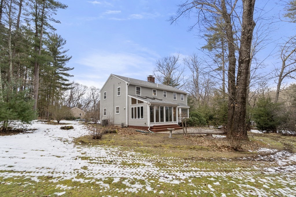 Two-story gray house with a glass sunroom and deck in a wooded yard with patchy snow.