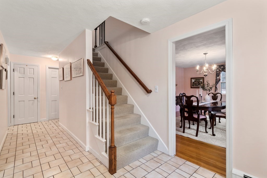 Entry hall with tiled floor and carpeted staircase, looking into a dining room with a chandelier.