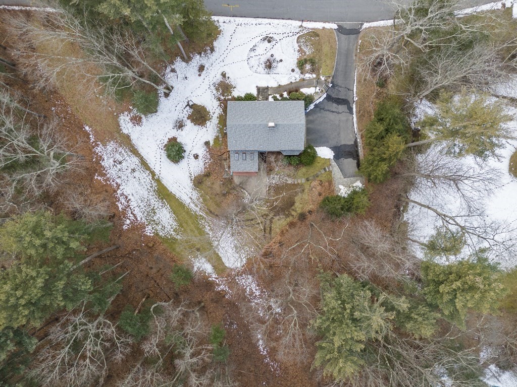Aerial view of a house surrounded by leafless trees, patchy snow, and a paved driveway.