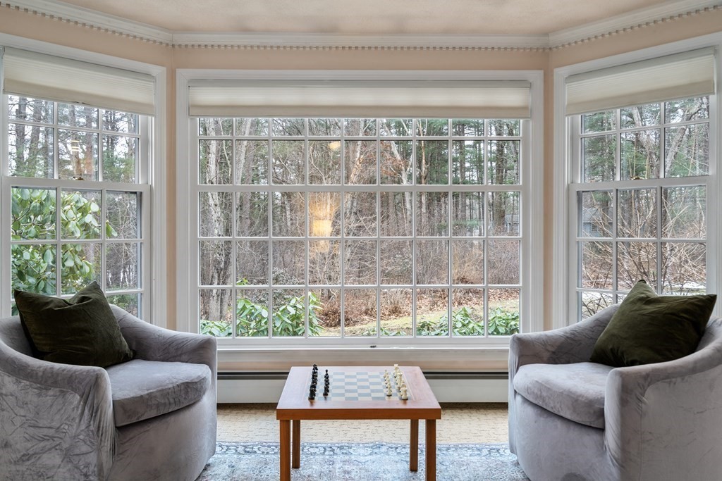 Sitting area with gray armchairs, a chess table, and a large bay window overlooking woods.