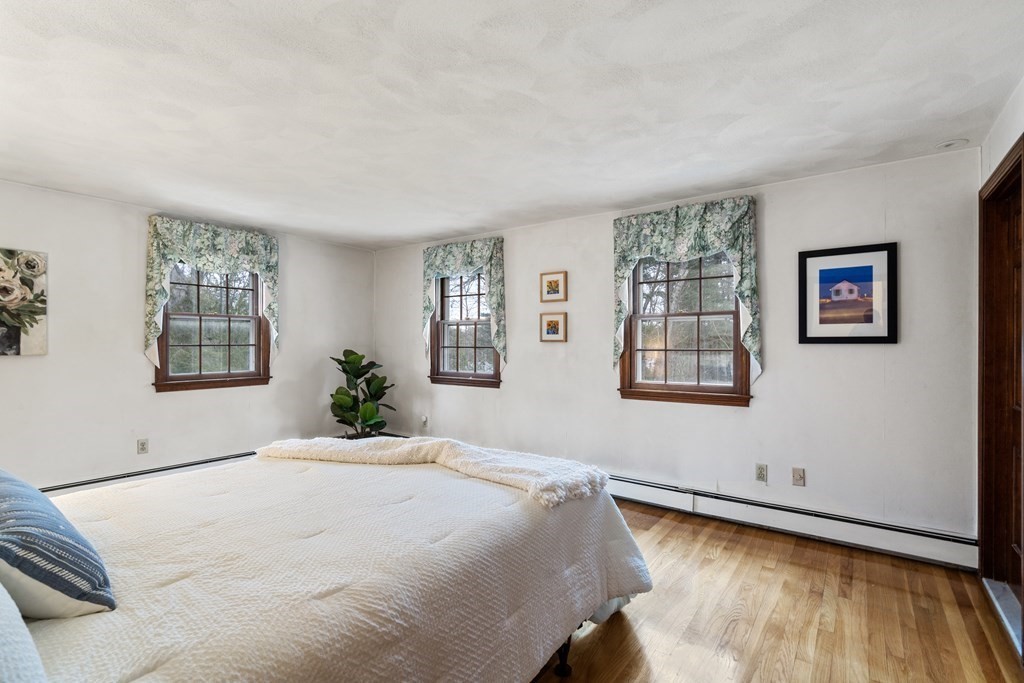 Bedroom with a large white bed, three curtained windows, and hardwood flooring.