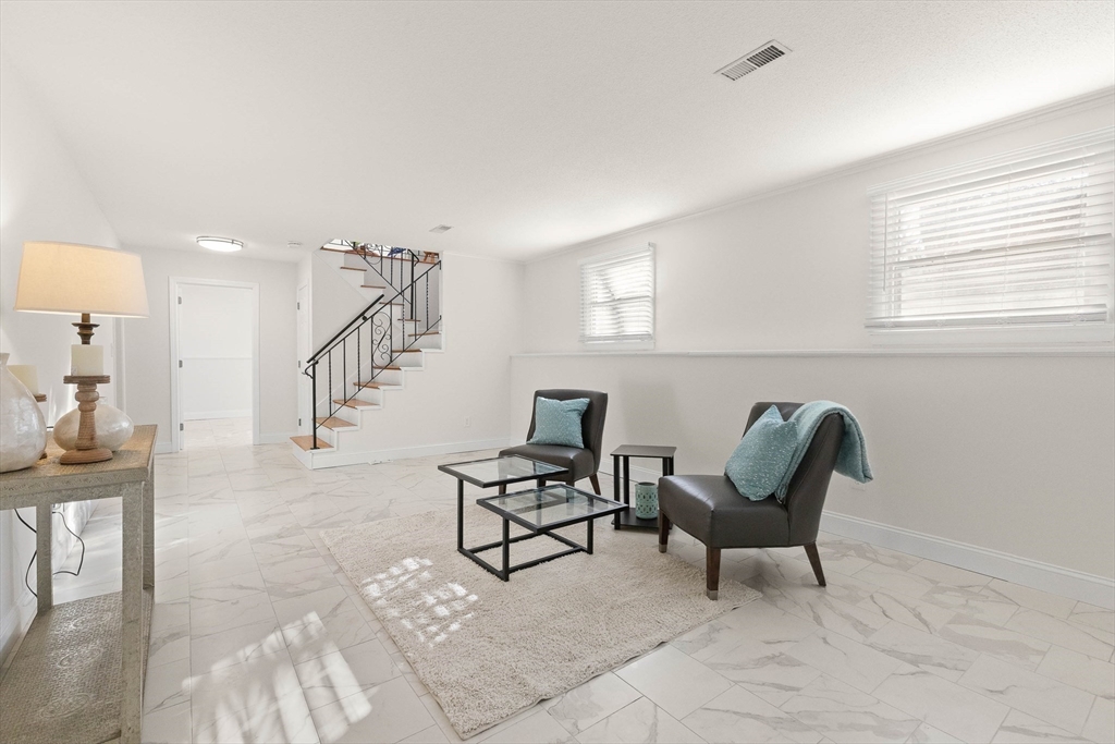 Furnished living room with marble-look tile floors, teal cushions, and a black-railed staircase.