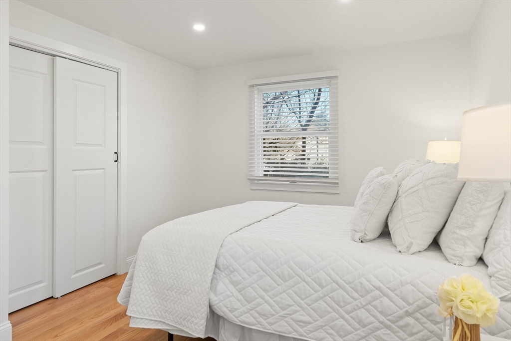 Bright bedroom with white quilted bedding, a window with blinds, and light wood flooring.