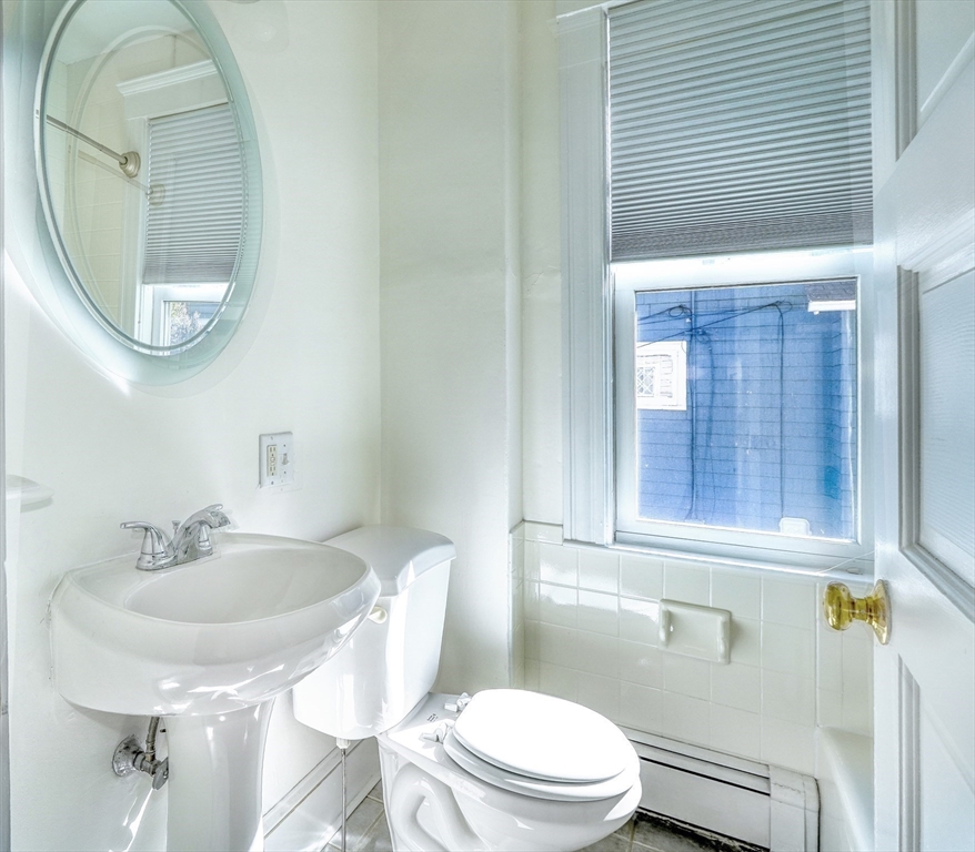 White bathroom with a pedestal sink, toilet, oval mirror, and window with a pleated blind.