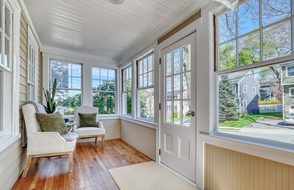 Enclosed sunroom with two upholstered chairs, wood floors, and large windows overlooking neighboring homes.