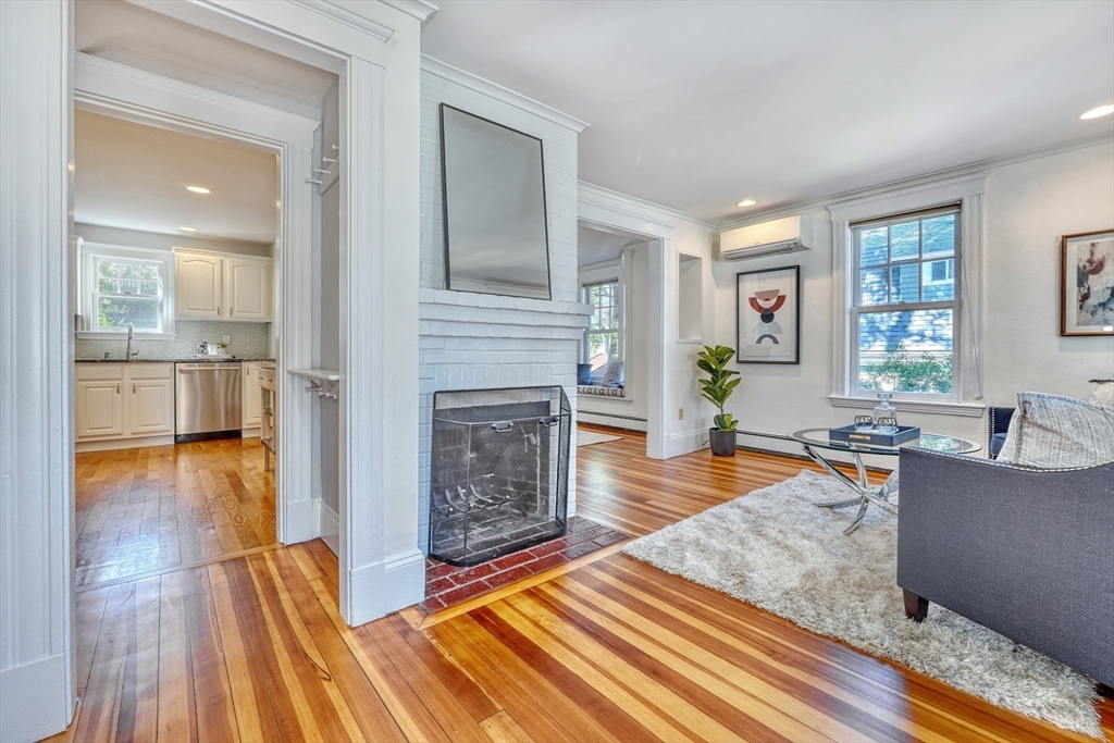 Living room with a white brick fireplace, striped hardwood floors, and an adjoining kitchen.
