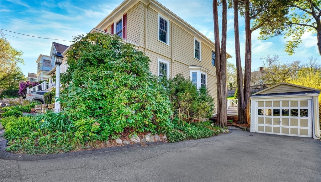 Beige house with detached garage, broad driveway, mature trees, and landscaped shrubs.