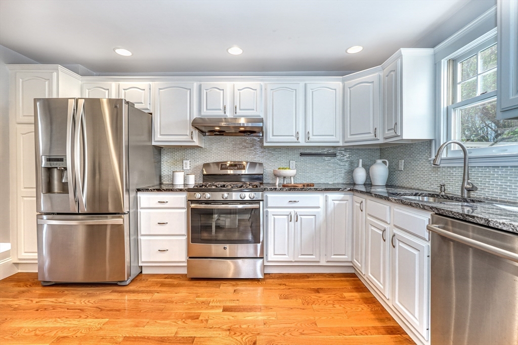 Kitchen with white cabinets, stainless steel appliances, gray tile backsplash, and warm hardwood floors.